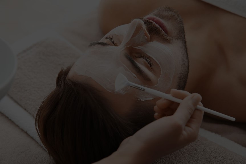 Person lying down with eyes closed, receiving a facial treatment with a brush applying cream.